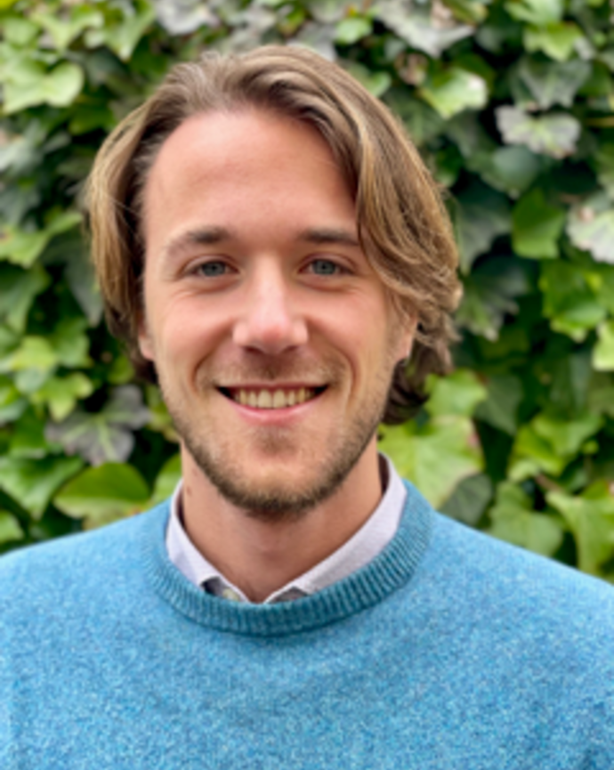 A person wearing a blue sweater and standing in front of foilage